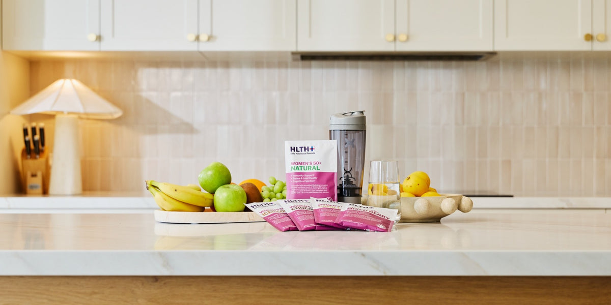 HLTH+ Women's 50+ Natural sachets on a marble kitchen bench with an electric shaker, a glass of water and fresh fruit — HLTH+