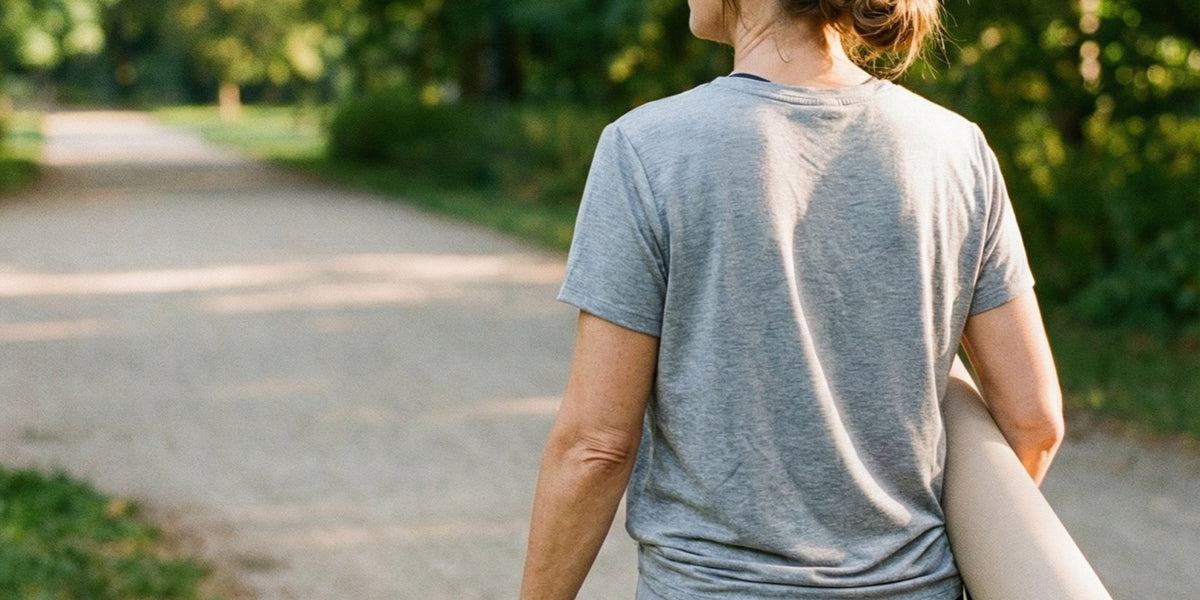A woman seen from behind jogging along a sunlit, tree-lined park path with a rolled yoga mat under her arm — HLTH+