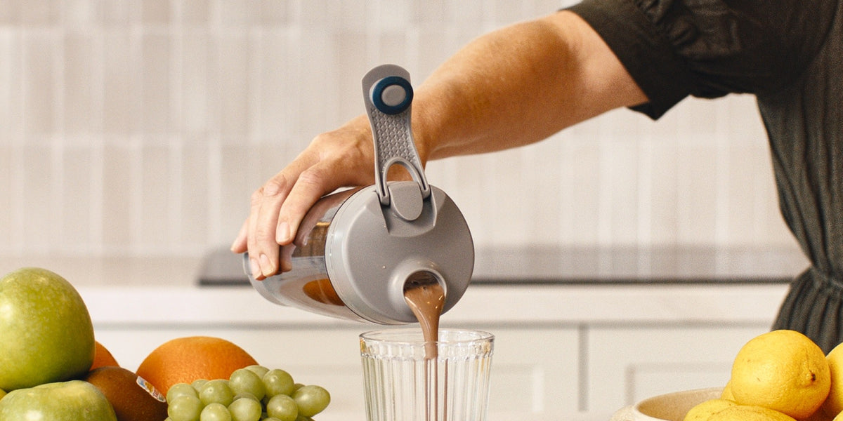 Hands pouring a chocolate-coloured nutritional formula from an electric shaker into a clear glass on a light wood kitchen bench — HLTH+