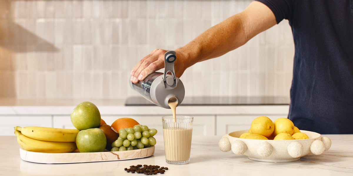A man in his fifties pouring a shake from a HLTH+ electric shaker into a glass in a bright tiled kitchen — HLTH+