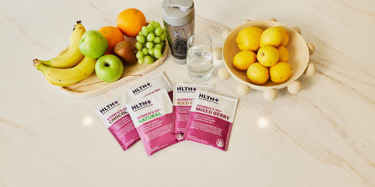 Three pink HLTH+ Women's sachets in Chocolate, Natural and Mixed Berry on a marble bench with fresh fruit, a charcoal shaker and a glass of water — HLTH+