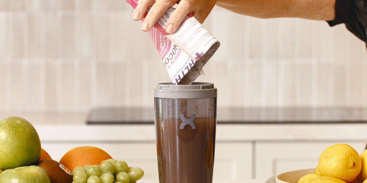 A woman's hand pours a pink Women's HLTH+ sachet into a charcoal electric shaker on a bright kitchen bench, surrounded by fresh fruit — HLTH+