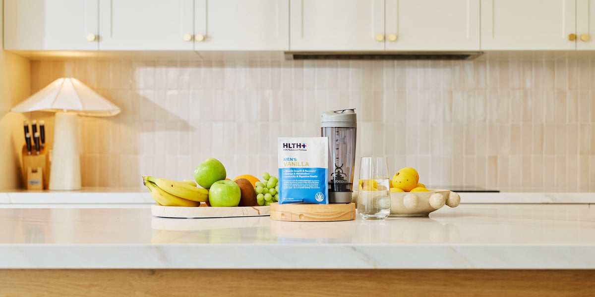 A HLTH+ Men's Vanilla sachet and electric shaker on a white marble kitchen bench with fresh fruit — HLTH+
