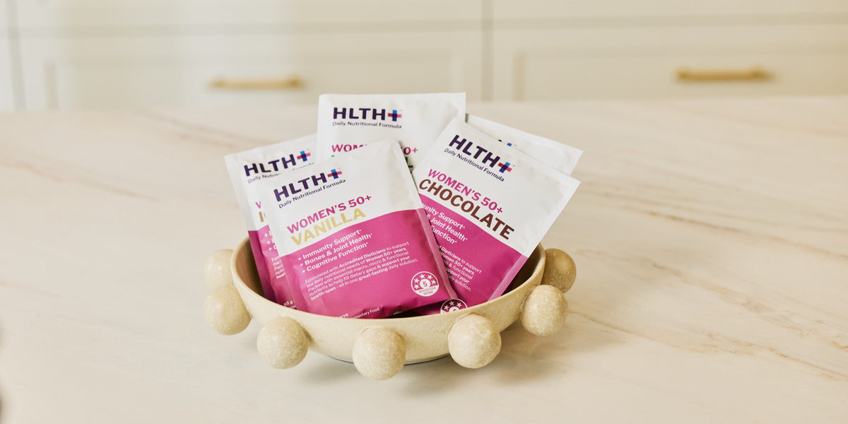 Three HLTH+ Women's 50+ sachets, Vanilla and Chocolate labels showing, arranged in a cream ceramic bowl on a pale wooden bench — HLTH+