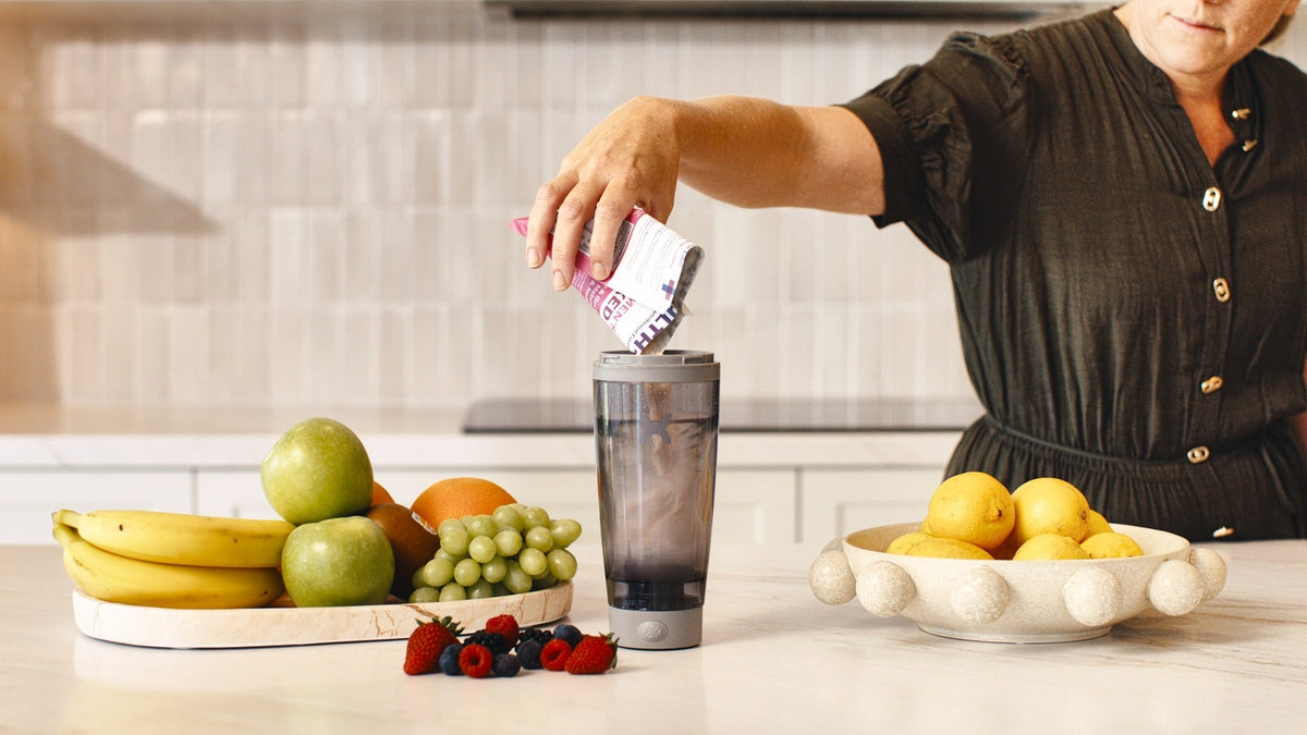 Woman pouring a HLTH+ Women's sachet into an electric shaker at a kitchen bench beside fresh fruit