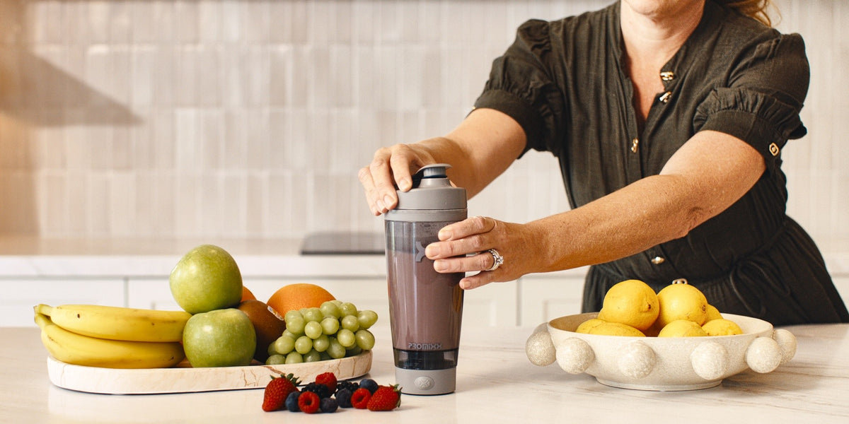 A woman in her fifties at a kitchen bench with her hands on a HLTH+ electric shaker, fresh produce on the boards beside her — HLTH+