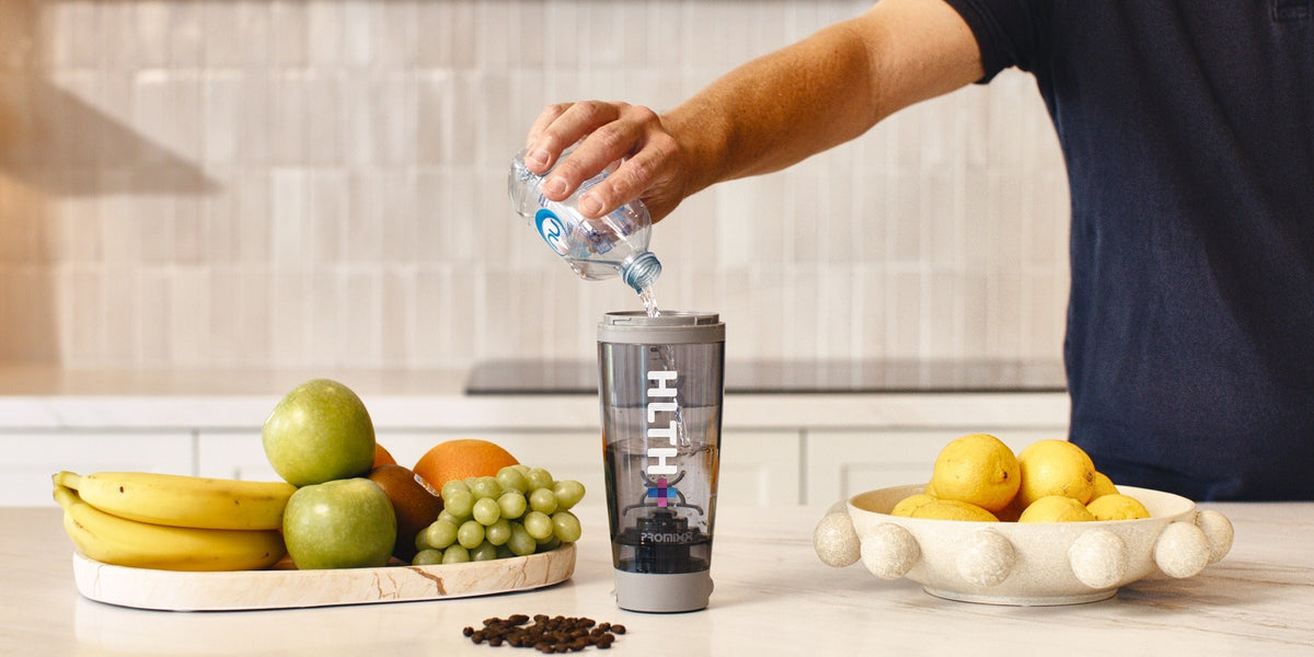 Hands pouring water from a glass into a HLTH+ electric shaker on a kitchen bench, fresh fruit in wooden bowls behind — HLTH+