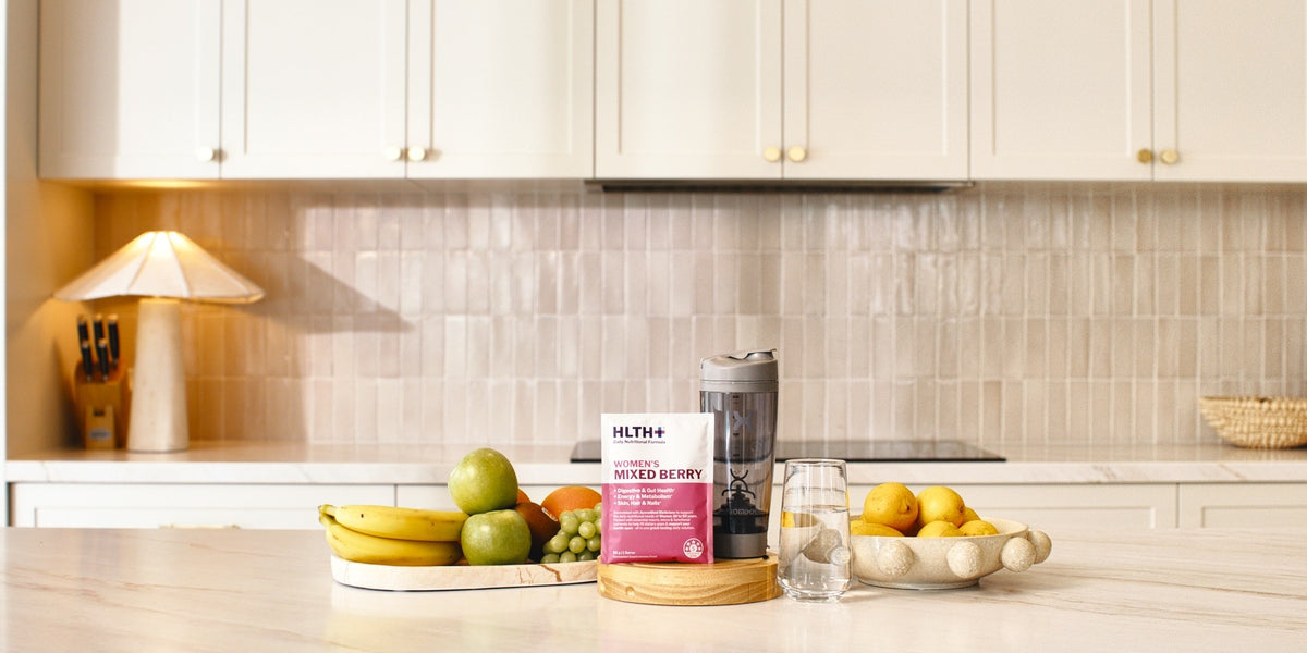 A Women's HLTH+ Mixed Berry sachet beside a charcoal electric shaker and a glass of water on a light wood kitchen bench, with fresh fruit in white bowls — HLTH+