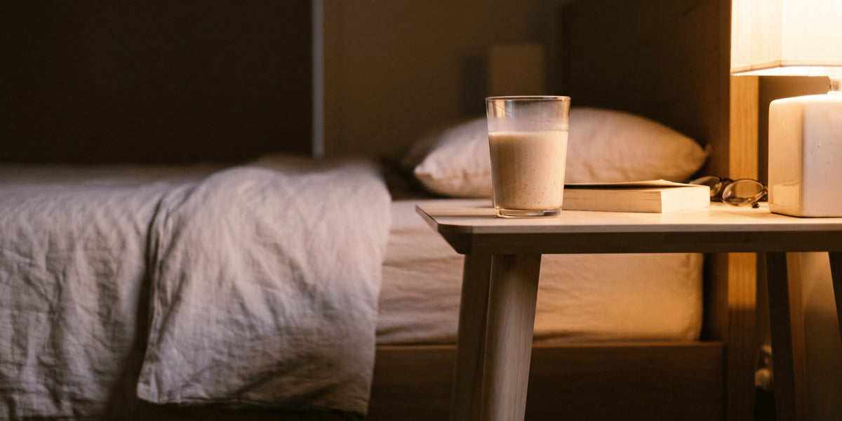 A glass of milky nutrition drink on a bedside table in warm lamplight — HLTH+