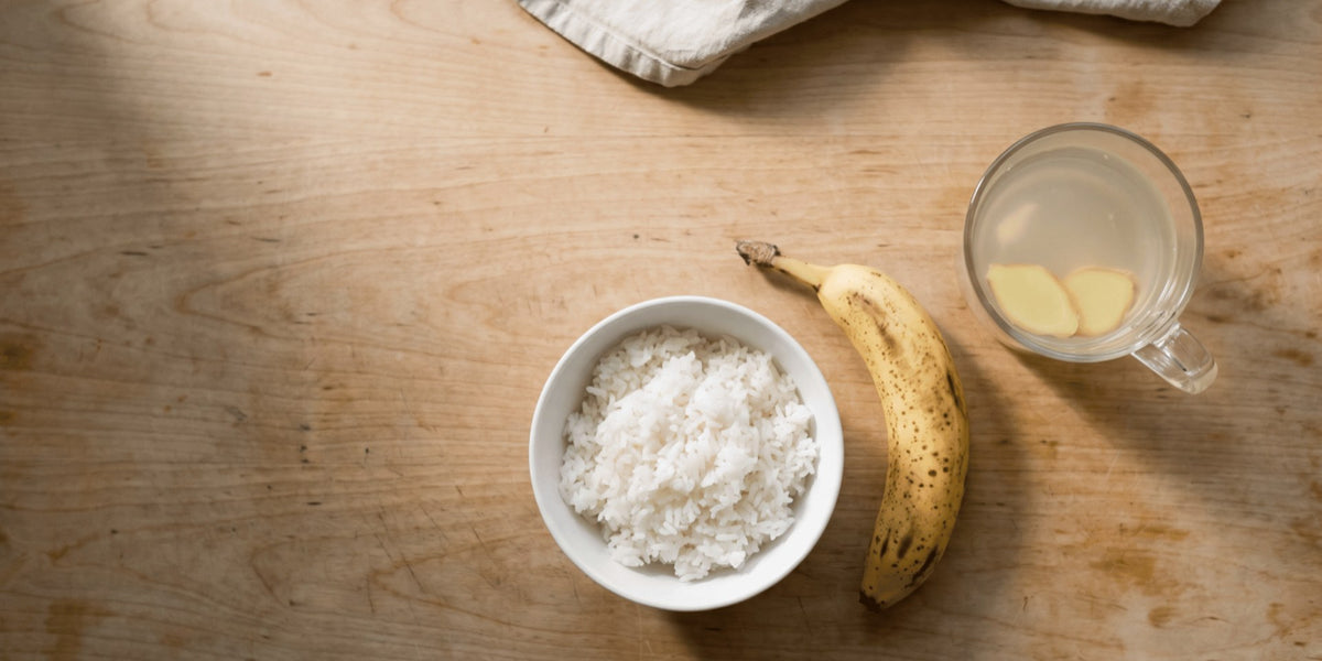 Overhead flat lay of a bowl of plain rice, a banana and a cup of ginger tea on a pale wooden bench — HLTH+