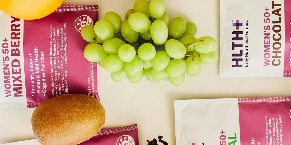 Four HLTH+ Women's 50+ sachets laid out with grapes, a green apple, an orange, a banana and a wholemeal bread roll — HLTH+