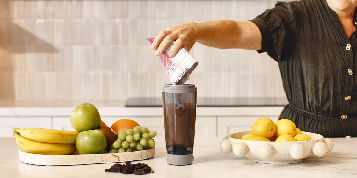 A woman pouring a pink HLTH+ Women's sachet into an electric shaker on a wooden kitchen bench beside fresh fruit — HLTH+