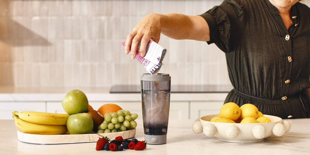 A woman in her 50s pours a pink HLTH+ sachet into a charcoal electric shaker on a bench surrounded by bananas, apples, grapes and berries — HLTH+