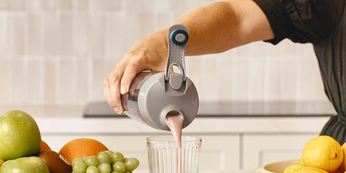 Hands in a black sleeve pour a pale pink drink from a charcoal electric shaker into a ribbed glass, with berries, apples and grapes around the bench — HLTH+