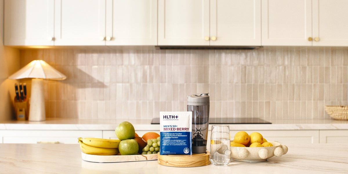 An HLTH+ Men's 50+ Mixed Berry sachet front of pack, centre stage on a light wood bench in a bright kitchen, beside a black electric shaker and a glass of water — HLTH+