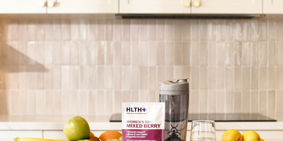 HLTH+ Women's 50+ Mixed Berry sachet on a wooden coaster with a black electric shaker and a glass of water on a wide kitchen bench — HLTH+