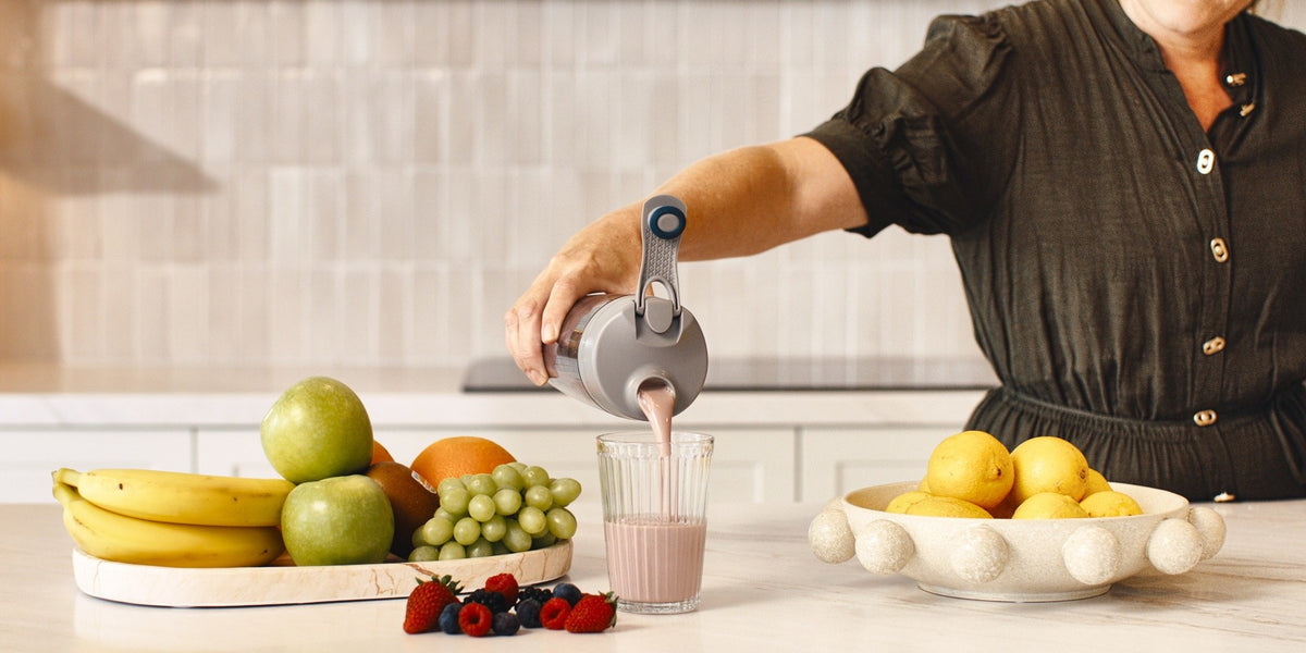 A woman in her fifties pouring a shake from a HLTH+ electric shaker into a glass on a bright kitchen bench with berries and fruit — HLTH+