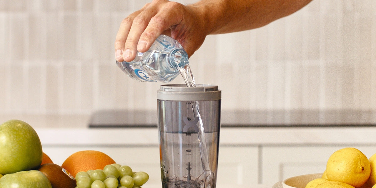 A hand pours water into a charcoal electric shaker on a light kitchen bench, with strawberries, blueberries, lemons and apples around it — HLTH+