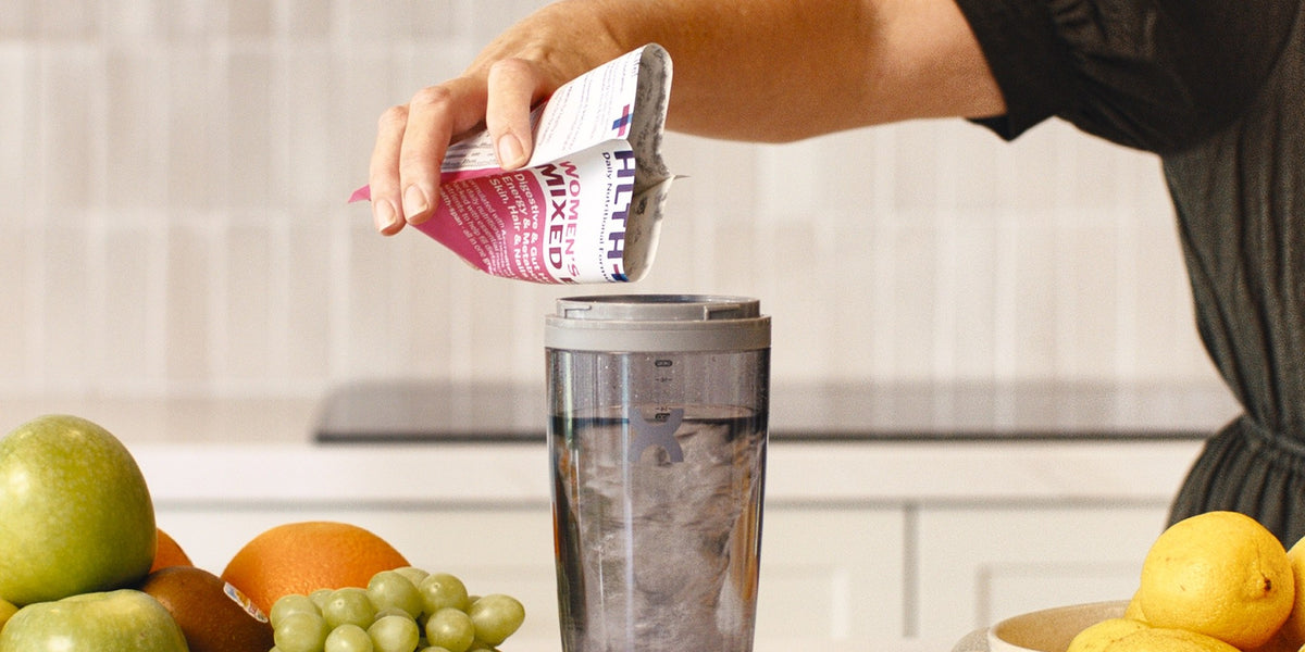 A woman in her 30s pours a pink Women's HLTH+ sachet into a charcoal electric shaker on a bright kitchen bench, with berries, apples and grapes around her — HLTH+