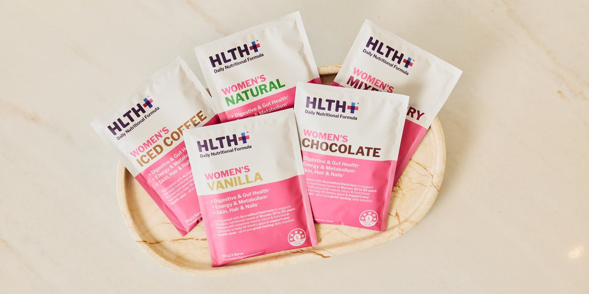 Five HLTH+ Women's Daily Nutritional Formula sachets in Natural, Mixed Berry, Iced Coffee, Vanilla and Chocolate on a round wooden tray — HLTH+