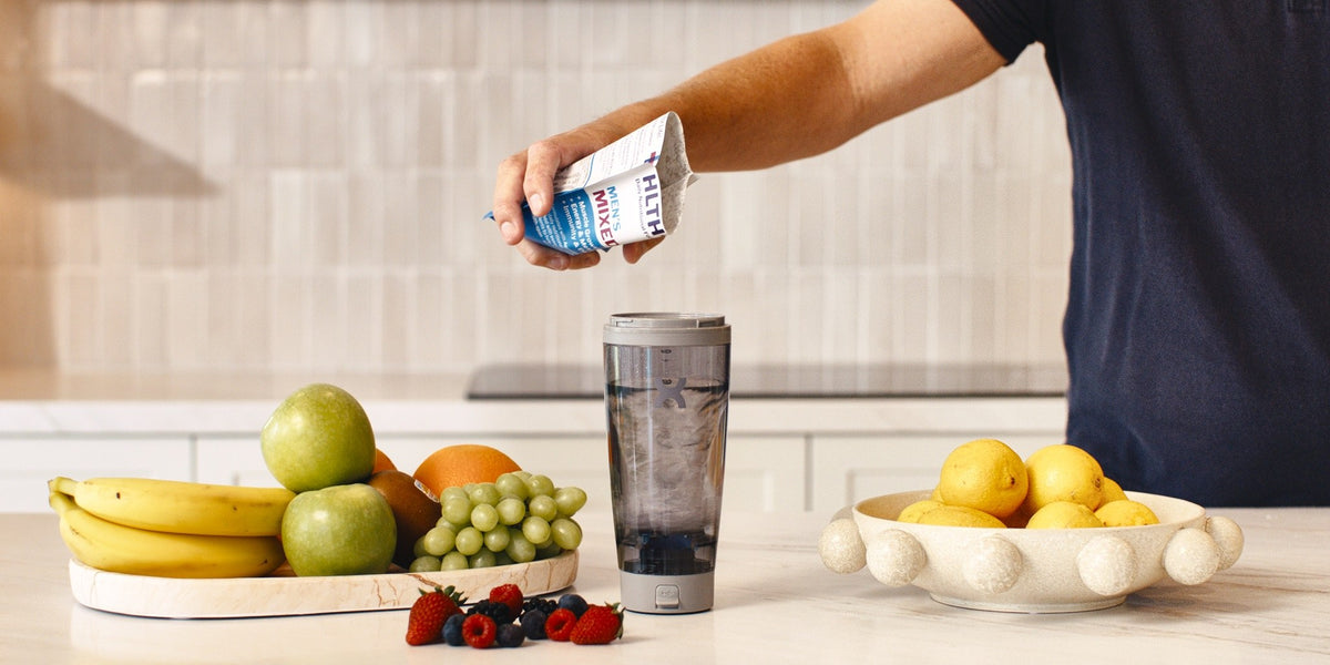 A hand pouring a HLTH+ sachet into a water-filled electric shaker on a wooden kitchen bench with fresh fruit — HLTH+
