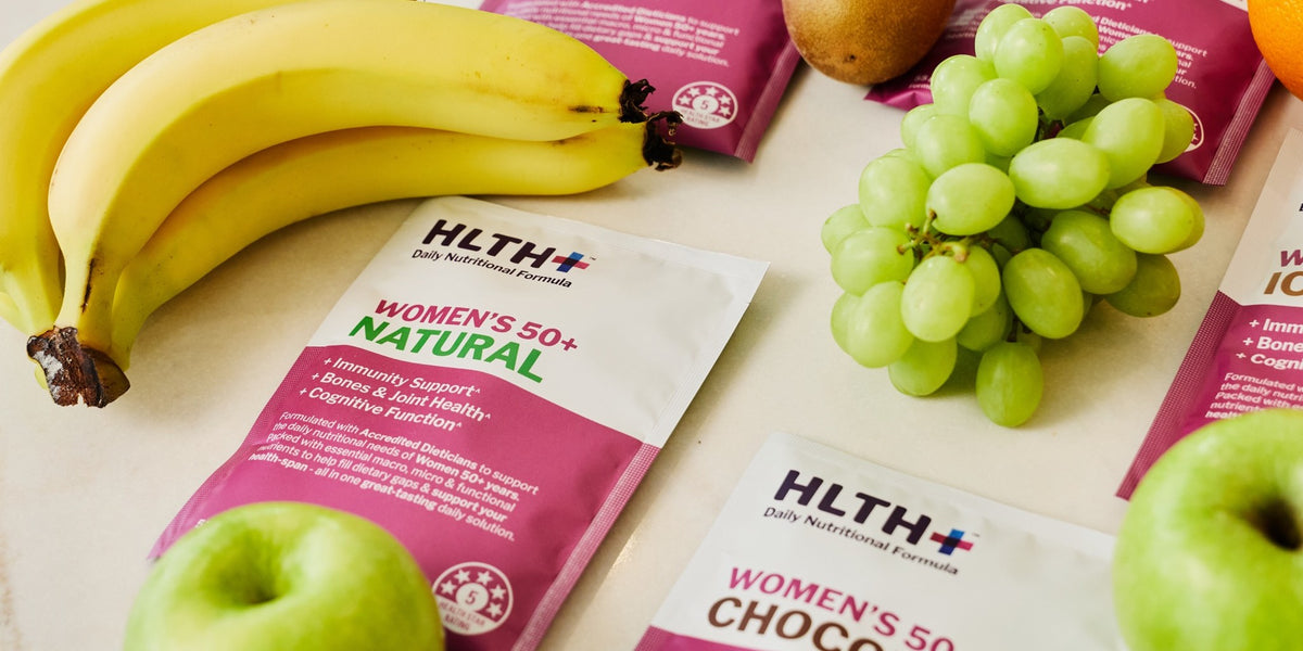 A studio flat lay of three HLTH+ sachets on a neutral beige surface surrounded by fresh fruit — HLTH+