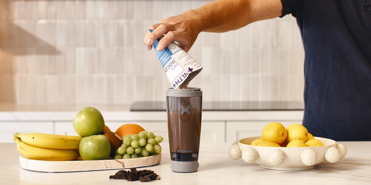 A hand pours a HLTH+ sachet into a charcoal electric shaker on a pale wood kitchen bench, fresh fruit and berries around it and bright window light behind — HLTH+