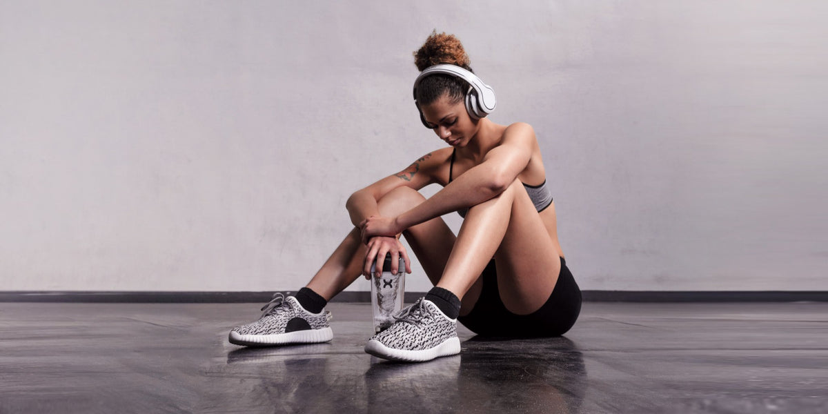 A woman in white headphones sits on a dark gym floor against a grey wall, holding a clear water bottle and looking down — HLTH+