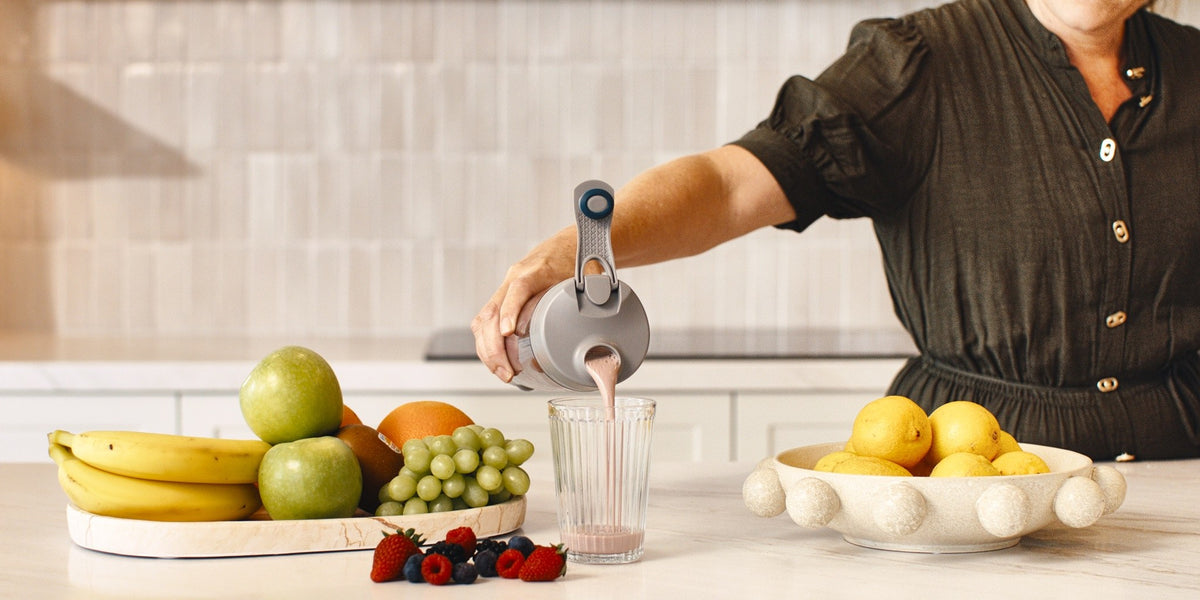 A woman in her fifties pouring a pink shake from a HLTH+ electric shaker into a glass on a white kitchen bench — HLTH+