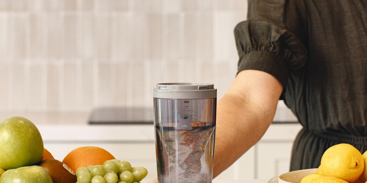 A woman in her 30s stands at a light wood kitchen bench holding an electric shaker of dark drink, with strawberries, blueberries, apples and grapes around her — HLTH+