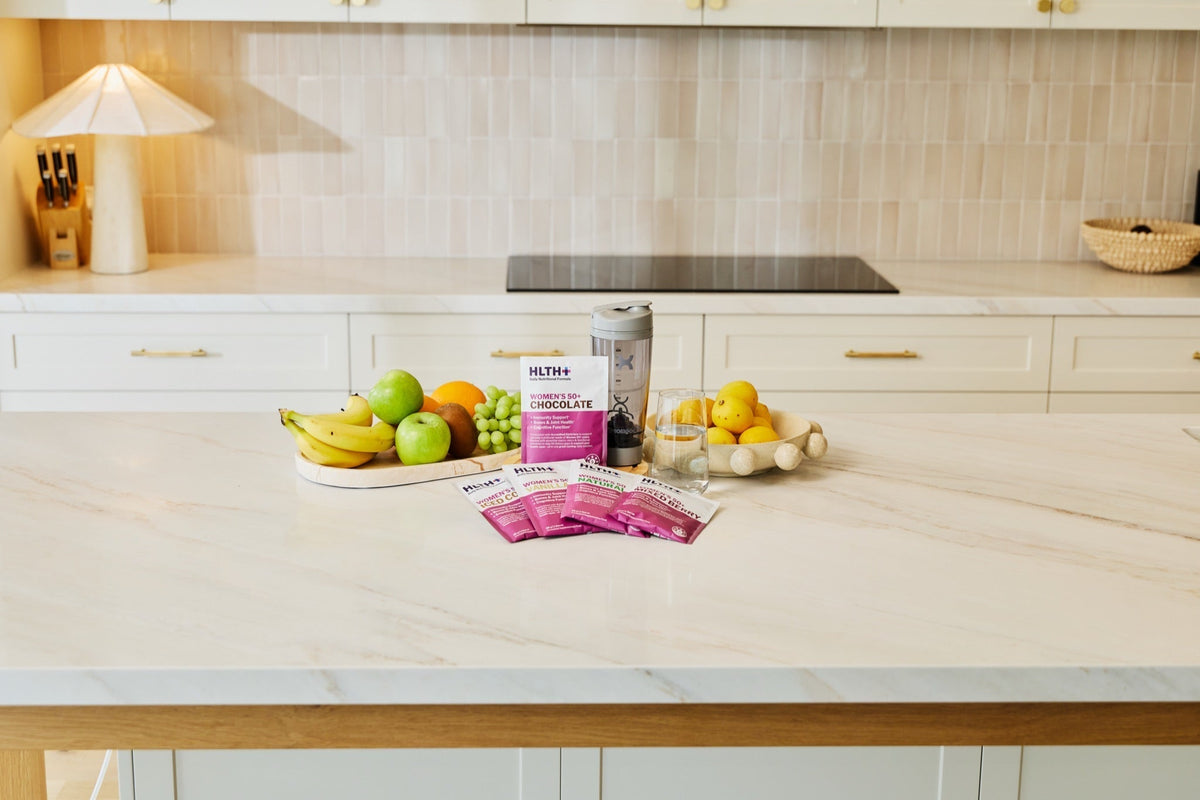 HLTH+ Women's 50+ sachets on a kitchen bench with fresh fruit, an electric shaker and a glass of water