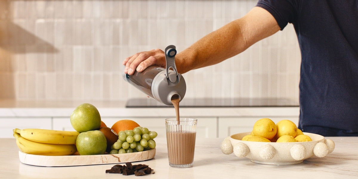 A man in his fifties pouring a shake from a HLTH+ electric shaker into a glass on a wooden kitchen bench — HLTH+