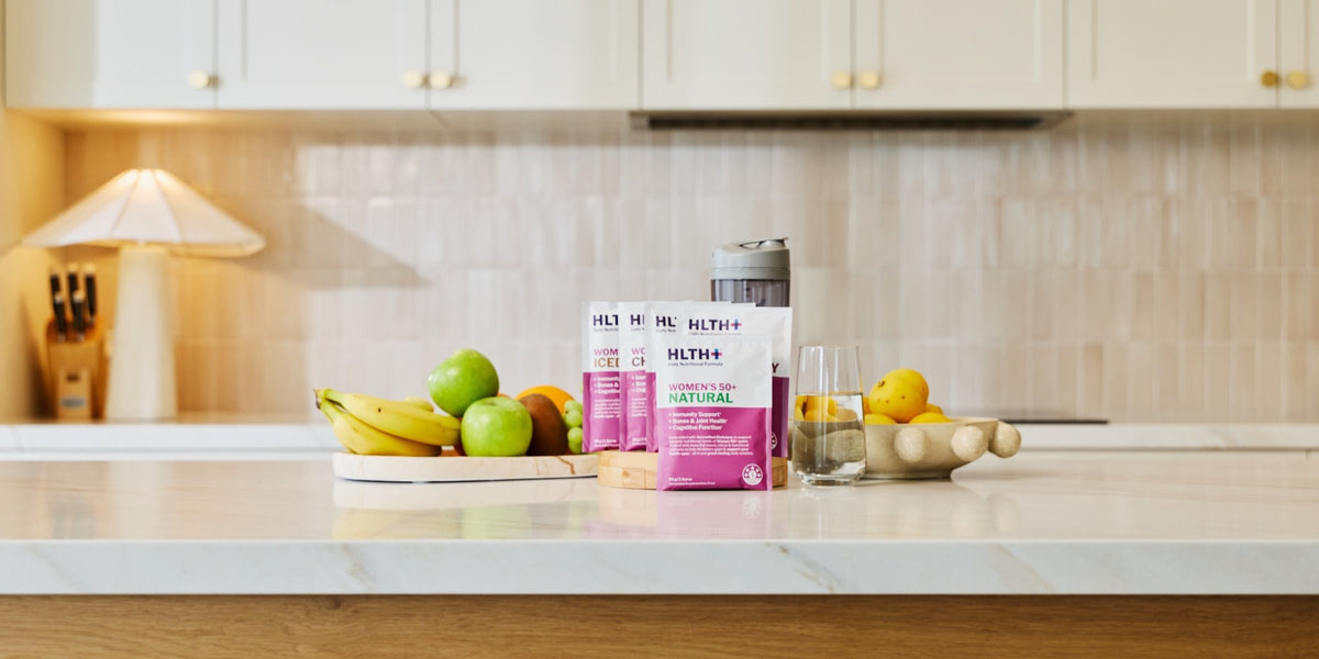 A HLTH+ Women's 50+ Natural sachet on a white marble bench styled with bananas, apples and a lemon — HLTH+