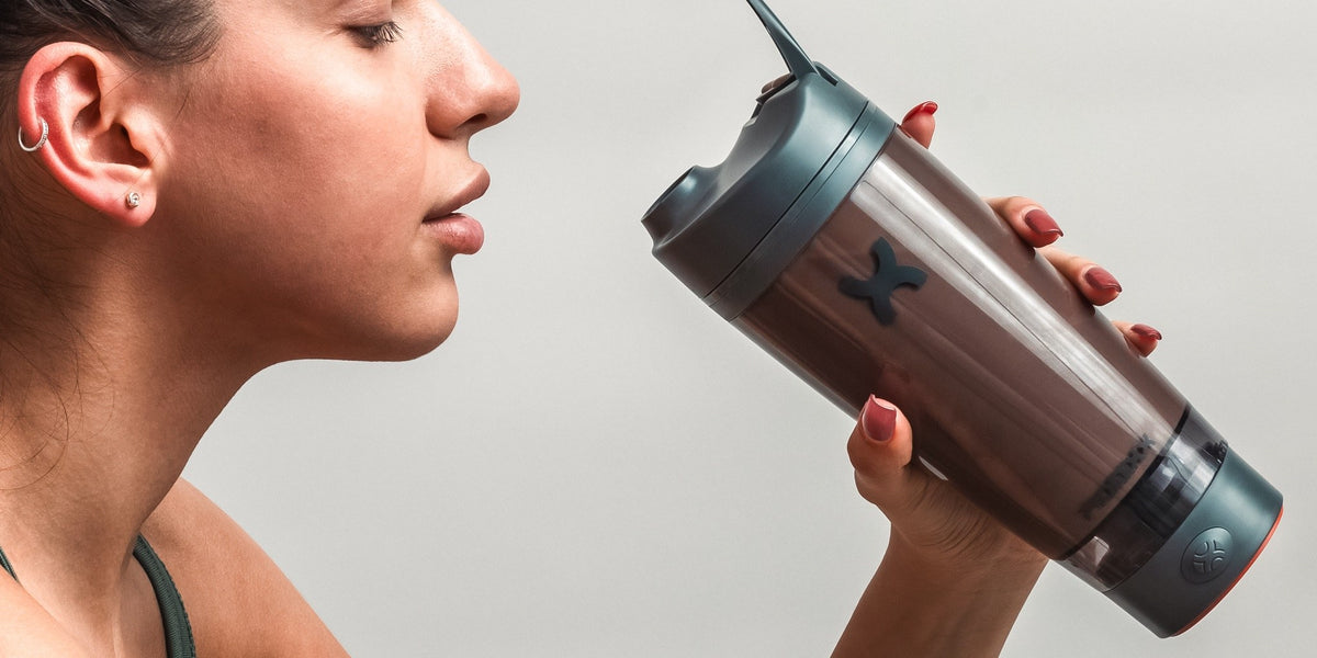 A woman in her 30s drinks from a charcoal electric shaker in profile against a soft grey studio backdrop — HLTH+