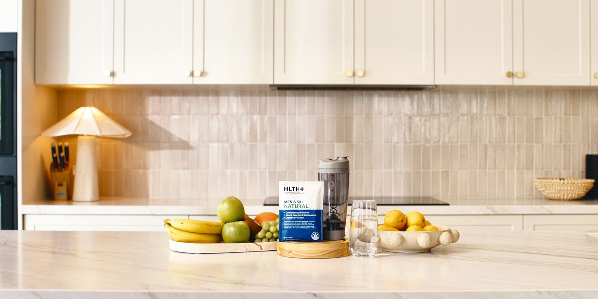 A HLTH+ Men's 50+ Natural sachet on a light marble benchtop with a black electric shaker and a glass of water — HLTH+
