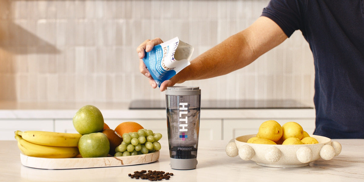 A man in his 30s pours an HLTH+ Men's sachet into a black electric shaker on a light wood bench, with fruit and coffee beans nearby — HLTH+