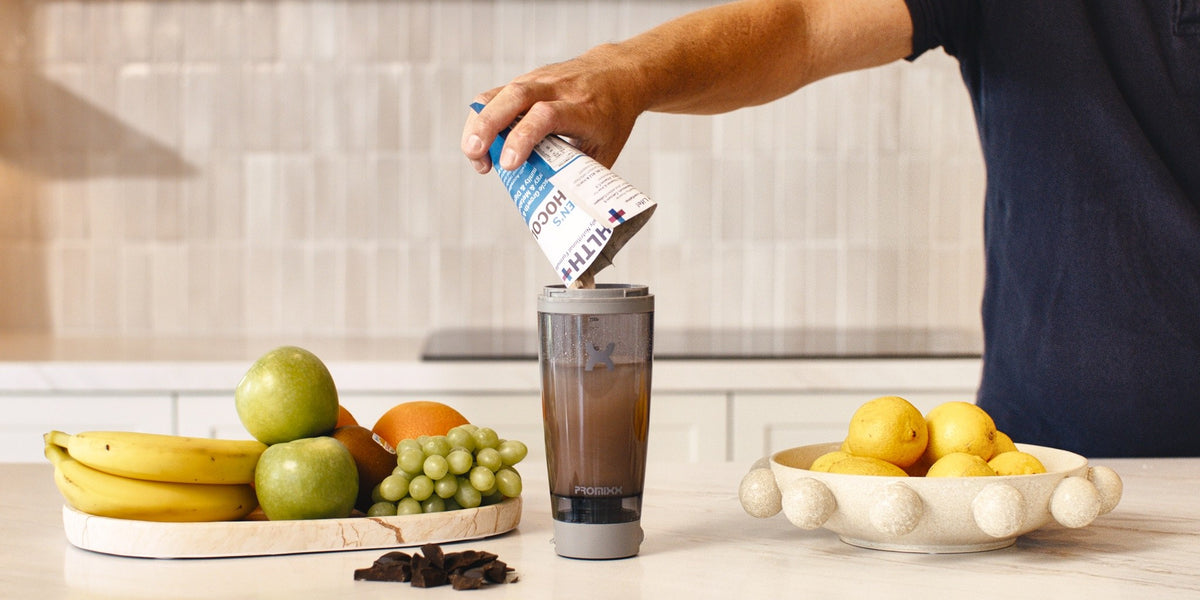 A hand pouring a HLTH+ Men's sachet into an electric shaker on a wooden kitchen bench — HLTH+