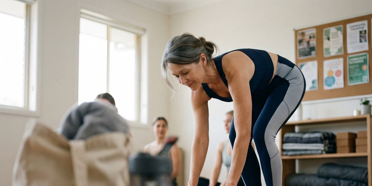 A woman in her fifties in athletic wear rolling up a yoga mat in a bright fitness studio — HLTH+