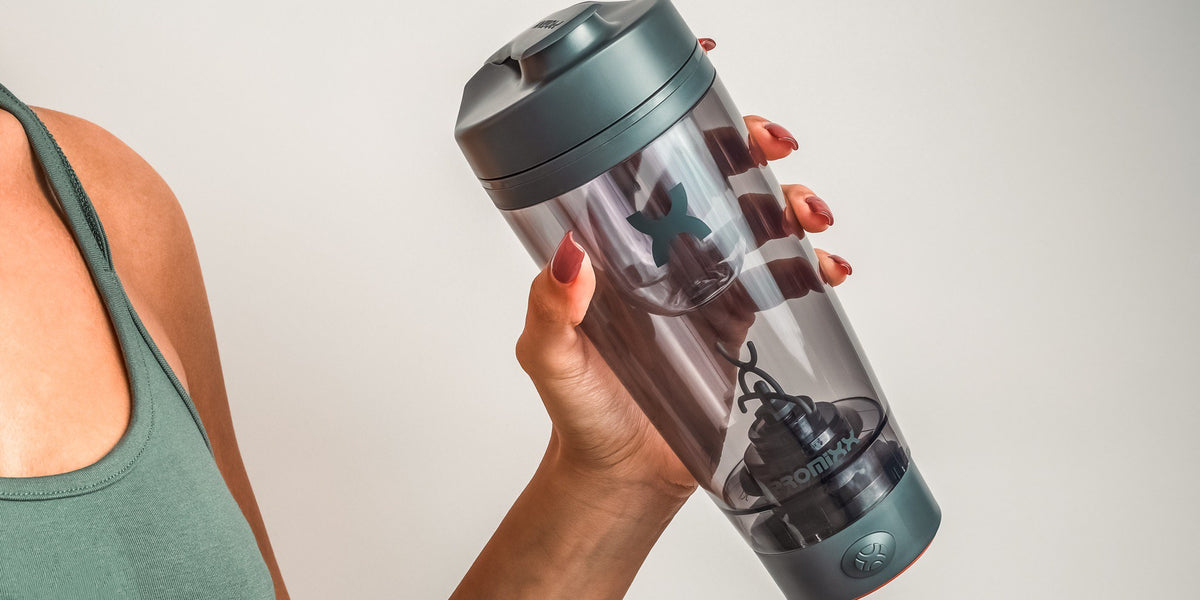 A woman in a sage-green sports top holds a charcoal and teal electric shaker in both hands against a plain white background — HLTH+