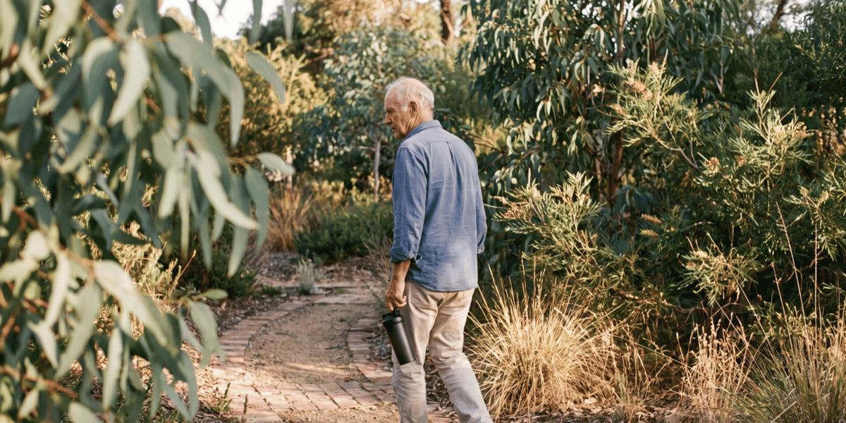 A man in his seventies walking along a garden path with a shaker in his hand — HLTH+
