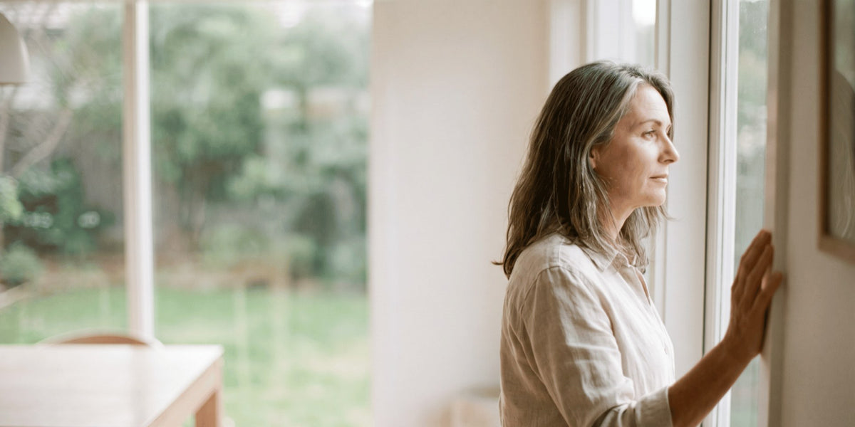 A woman around fifty standing at a kitchen window looking out at the garden — HLTH+