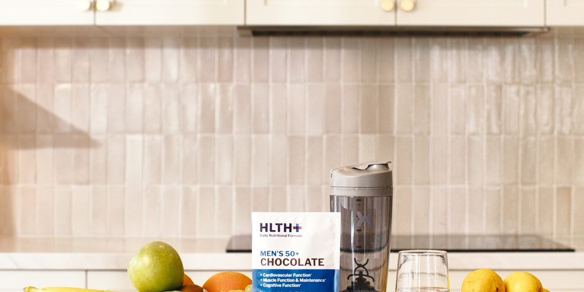 HLTH+ Men's 50+ Chocolate sachet on a wooden coaster beside a black electric shaker, with bowls of fruit and a glass of water on a marble bench — HLTH+