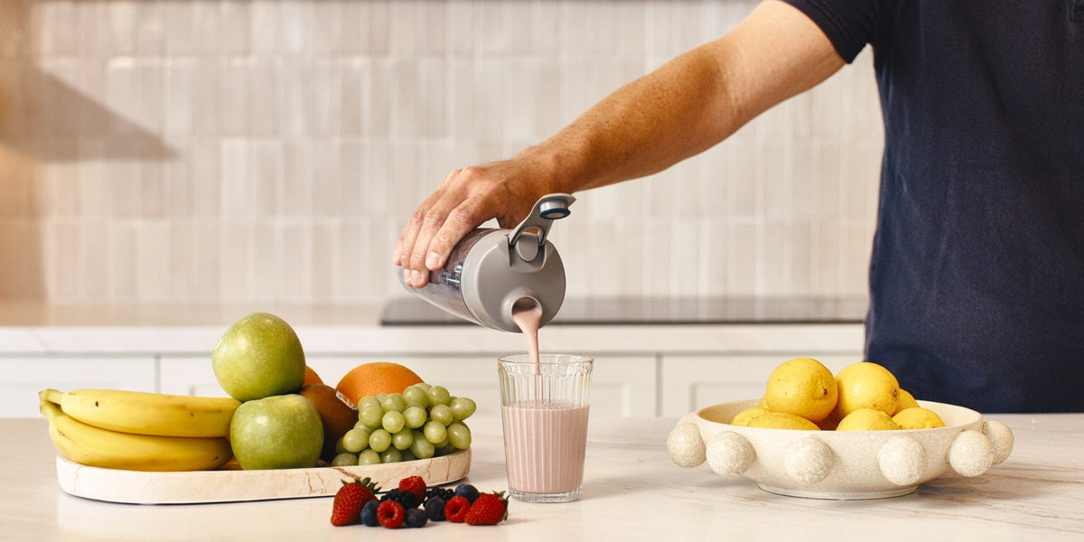 A man pouring a pale pink shake from a HLTH+ electric shaker into a glass on a wooden kitchen bench, fruit around the glass — HLTH+