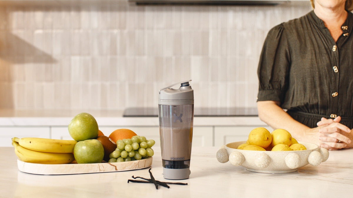 Woman standing calmly at a kitchen bench with a prepared HLTH+ shake and a tray of fresh fruit