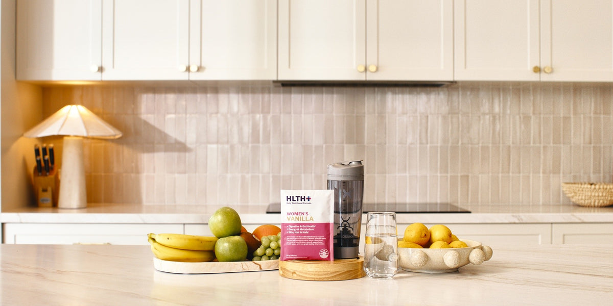 A HLTH+ Women's sachet on a light wood kitchen bench flanked by bananas, apples and grapes — HLTH+