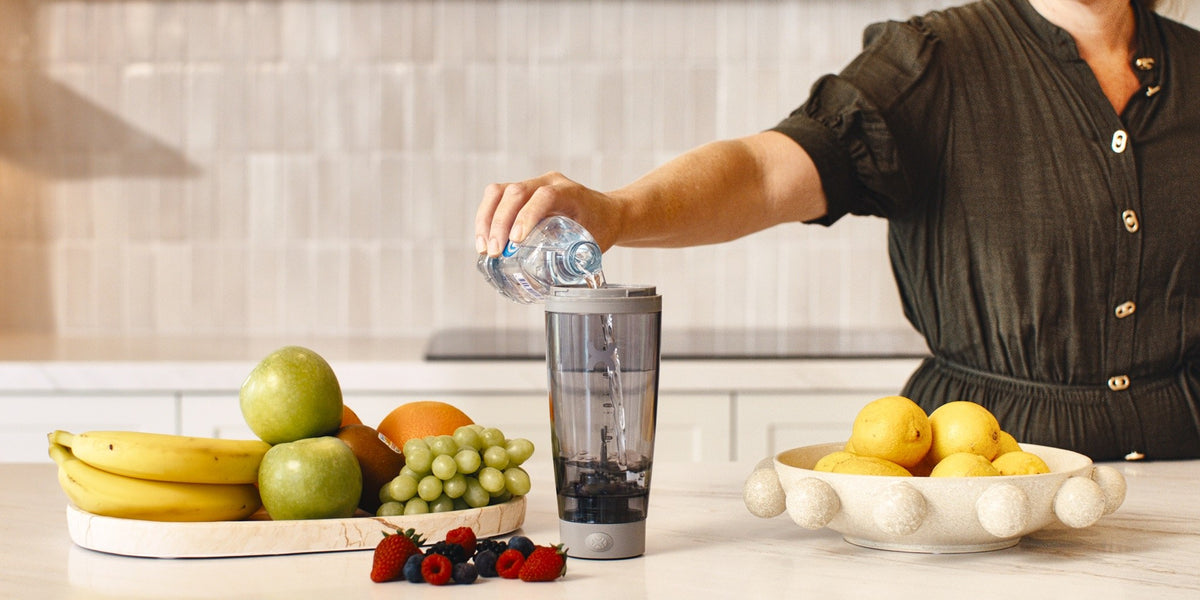 A woman pouring water into a blender on a wooden kitchen bench surrounded by bananas, apples, berries and lemons — HLTH+