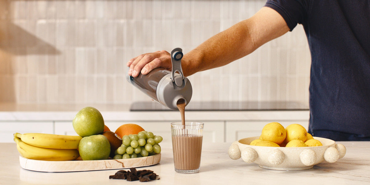 A man in a navy t-shirt pours a shake from a black HLTH+ electric shaker into a clear glass on a light wood bench, fresh fruit on wooden boards beside him — HLTH+