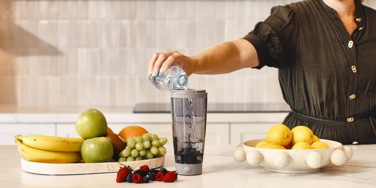A woman pouring water into a blender on a wooden kitchen bench with fresh fruit arranged beside it — HLTH+
