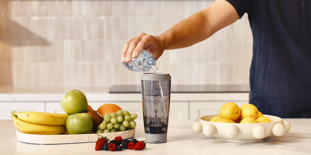 A man in his 30s pours water into a portable blender on a light wood kitchen bench, surrounded by bananas, berries, apples and lemons — HLTH+