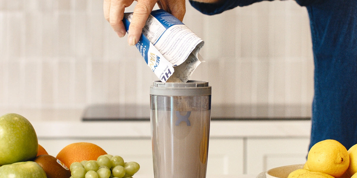 A man in his 50s pours a blue HLTH+ Men's 50+ sachet into a stainless steel electric shaker on a kitchen bench with fruit and eggs — HLTH+